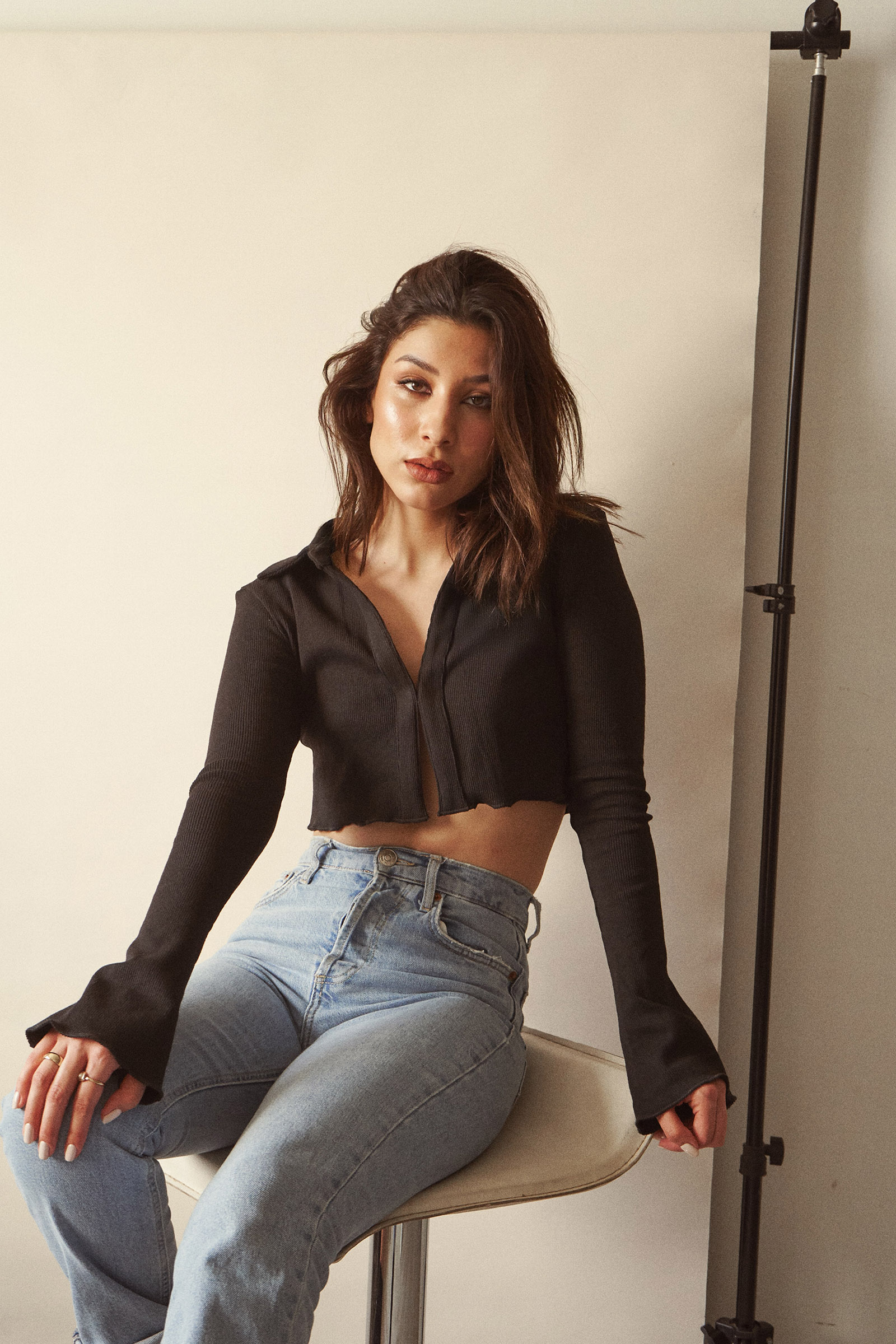 Nadya Inzunza is shown in a studio setting wearing a black crop top and blue jeans seated on a white stool against a beige backdrop.